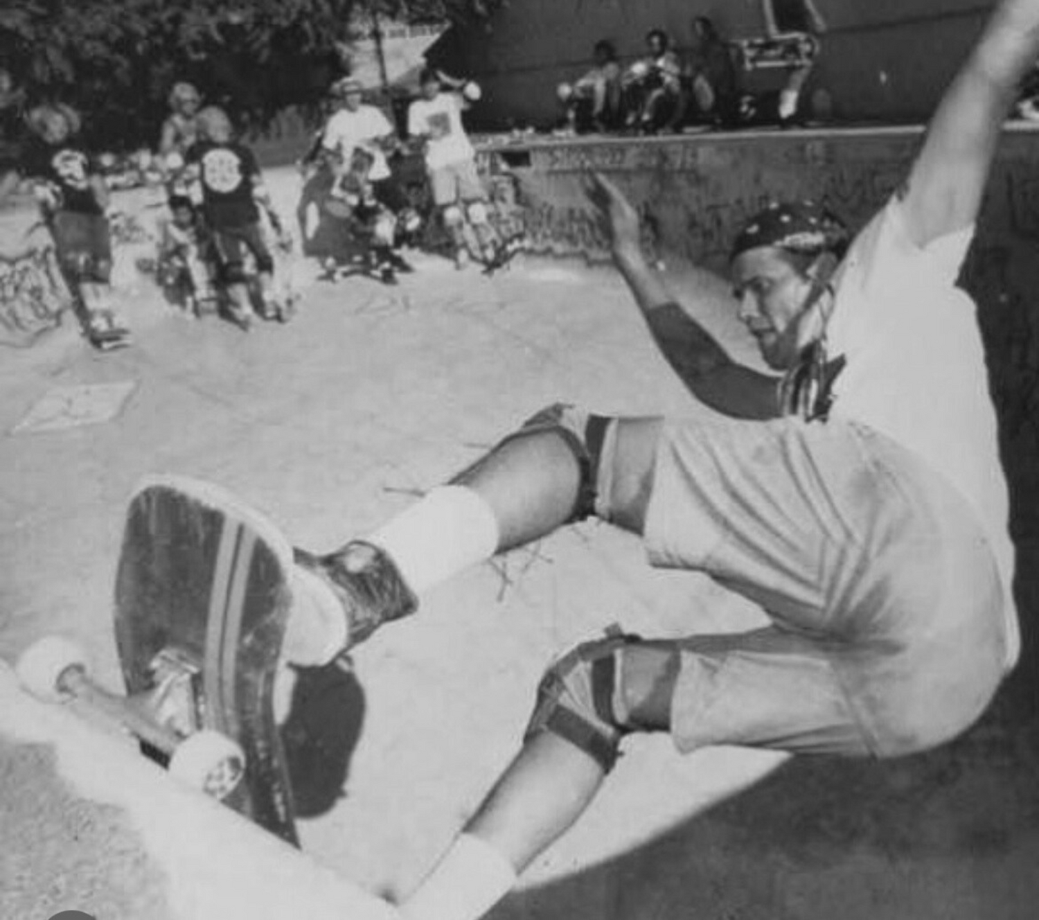 Ron Emory slides along pool coping with skaters gathered on the deck in the background.